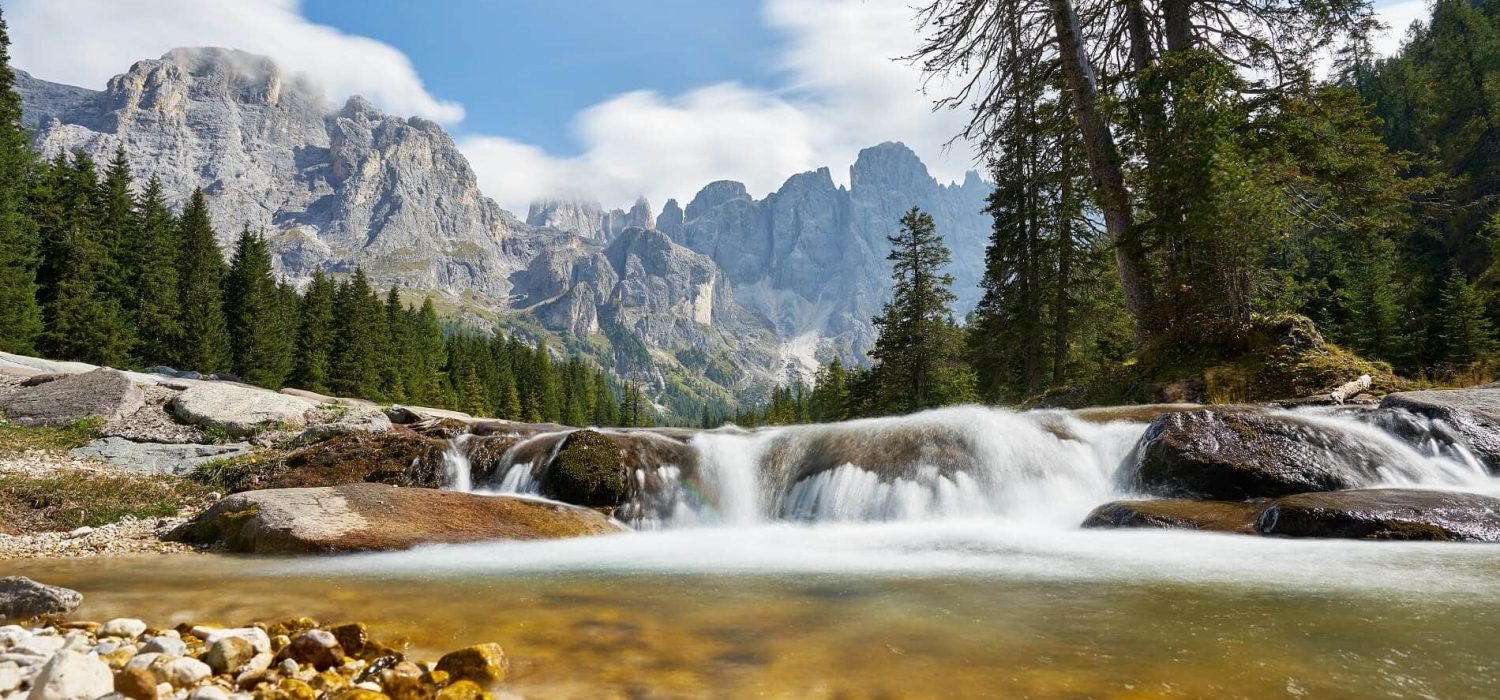 Ruscello montano che scorre tra alberi e montagne sotto un cielo sereno.